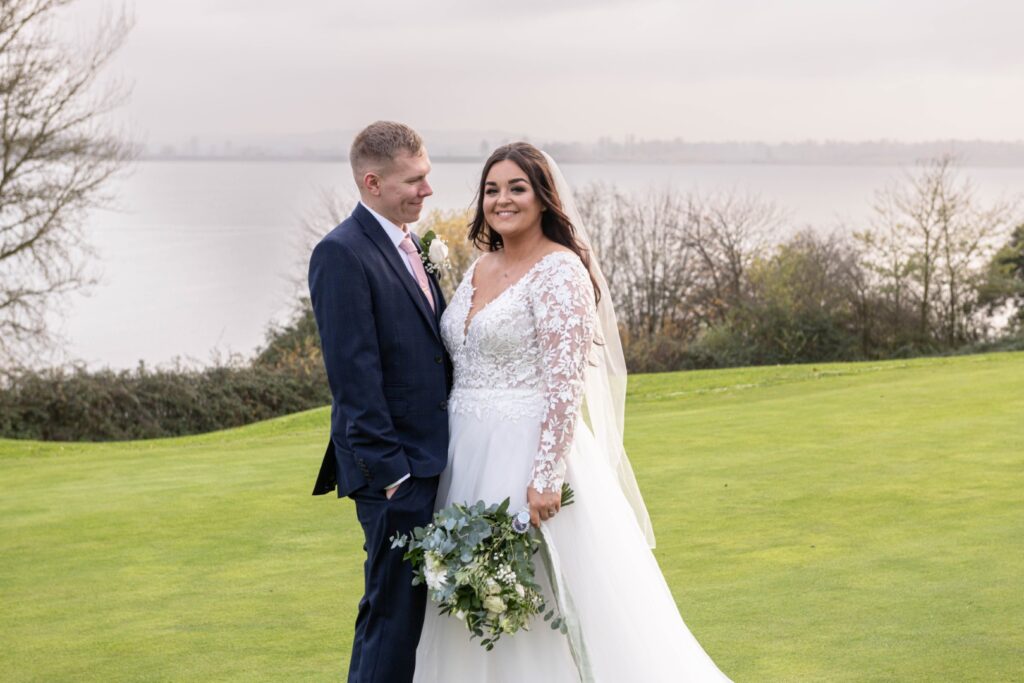 A happy couple poses together on the lush green golf course at Draycote Hotel by a serene water backdrop, showcasing the bride's beautiful floral lace gown and the groom's navy suit.