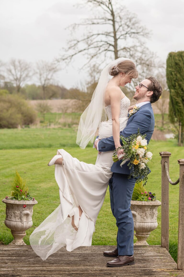 A bride and groom sharing a romantic moment outdoors, with the groom lifting the bride off the ground, as they gaze into each other's eyes and smile, surrounded by a pastoral landscape.