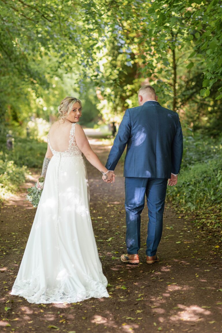 A bride in a white dress and a groom in a blue suit walking hand in hand along a tree-lined path, with the bride looking back over her shoulder towards the camera.