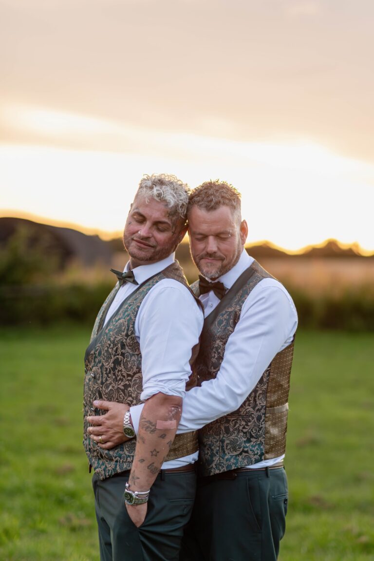 Two men in semi-formal attire with patterned vests and bow ties embracing and smiling against a sunset lit sky.