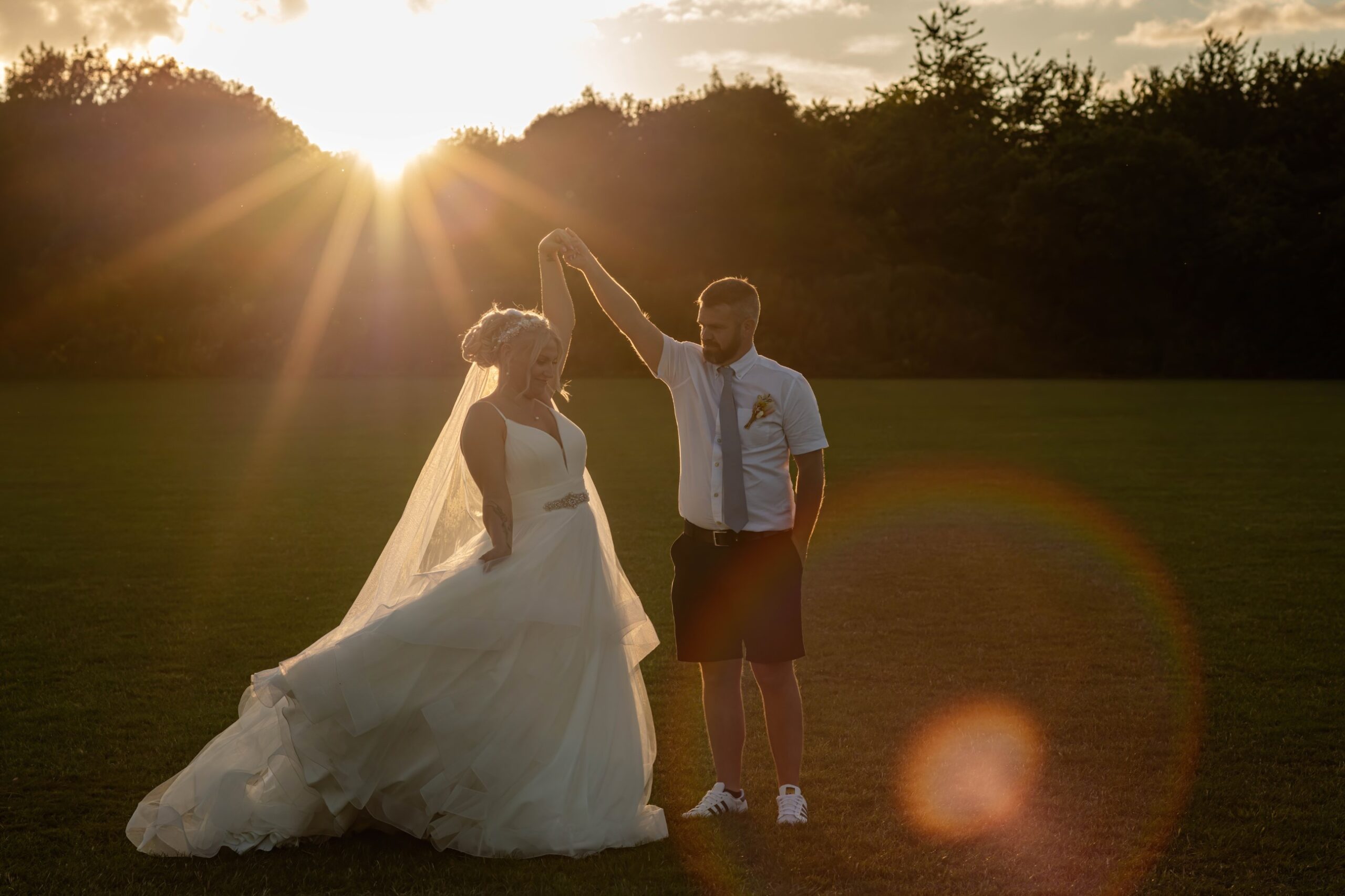 A bride in a white wedding dress and a groom in a white shirt and black shorts stand in a field with the sun setting in the background, creating a warm backlight and a slight lens flare. The groom is holding the bride's hand aloft as she looks down and smiles.