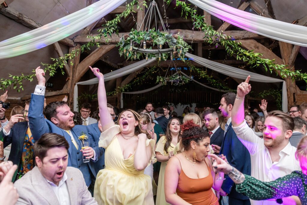 A group of guests dancing and celebrating enthusiastically at a wedding reception inside a rustic venue with wooden beams and fairy lights.