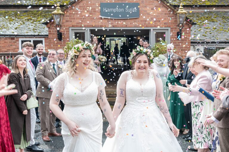 Two brides exiting the Hampton Suite building while guests throw confetti, celebrating the wedding with smiles and laughter.