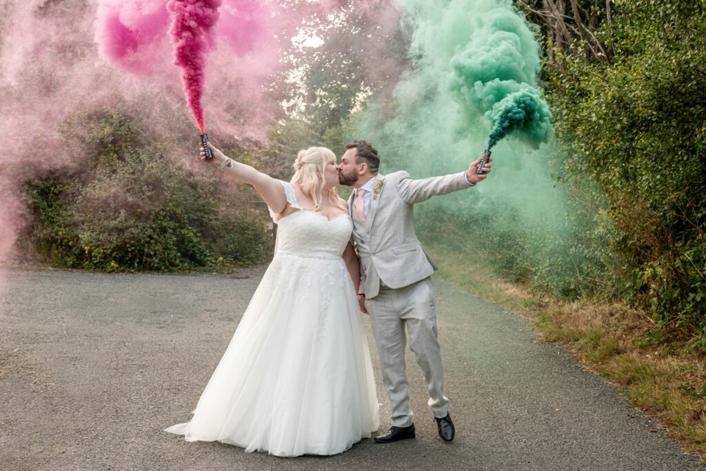 A bride and groom kissing while holding smoke bombs that release pink and green smoke, creating a vibrant backdrop on a path with greenery.