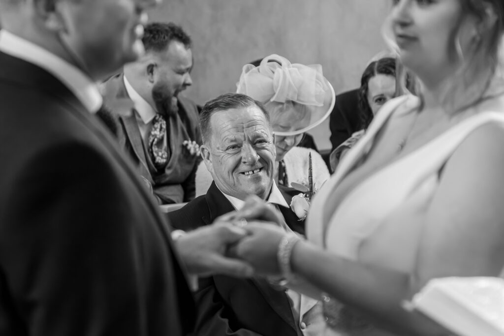 A father smiling brightly as watches his daughter getting married, with other guests in the background