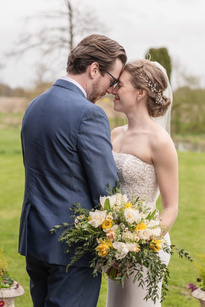 A bride and groom affectionately touching foreheads while holding a bouquet, with a soft-focus nature background.