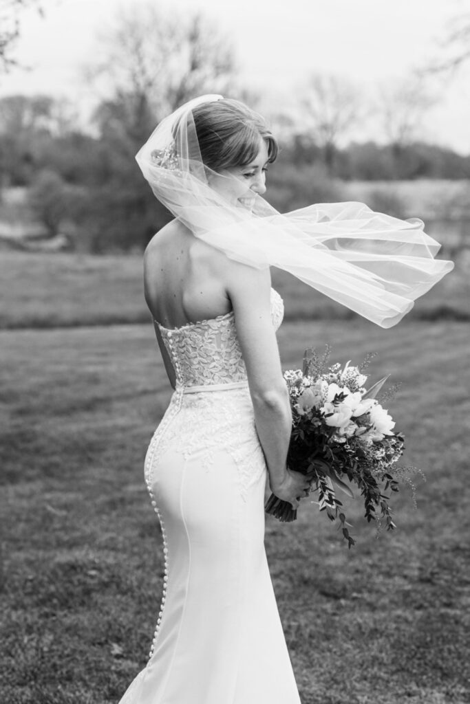 A black and white photo of a smiling bride with her veil flowing in the wind, holding a bouquet.