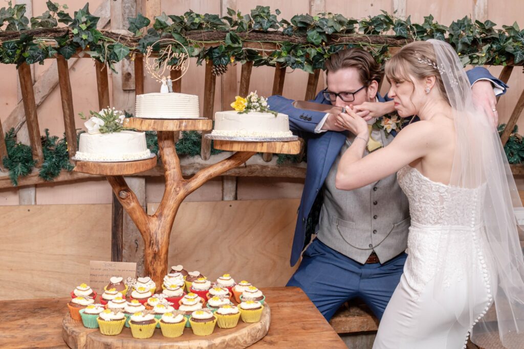 A bride and groom playfully preparing to cut their wedding cake, surrounded by cupcakes and rustic decorations.