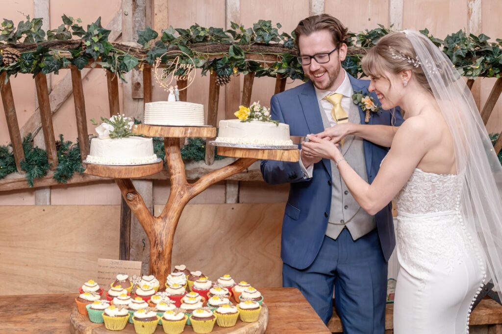 Bride and groom in wedding attire smiling and cutting a cake together at their wedding reception, with cupcakes and a personalized cake topper in the background