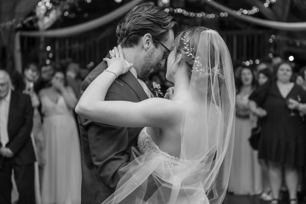 Black and white photo of a bride and groom sharing their first dance while guests watch in the background.