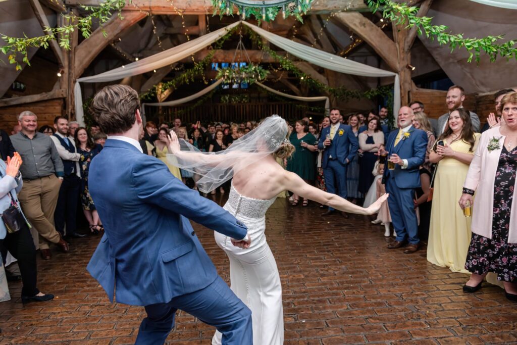 A bride and groom dancing enthusiastically in front of wedding guests at a rustic venue with the bride twirling her veil in the air.