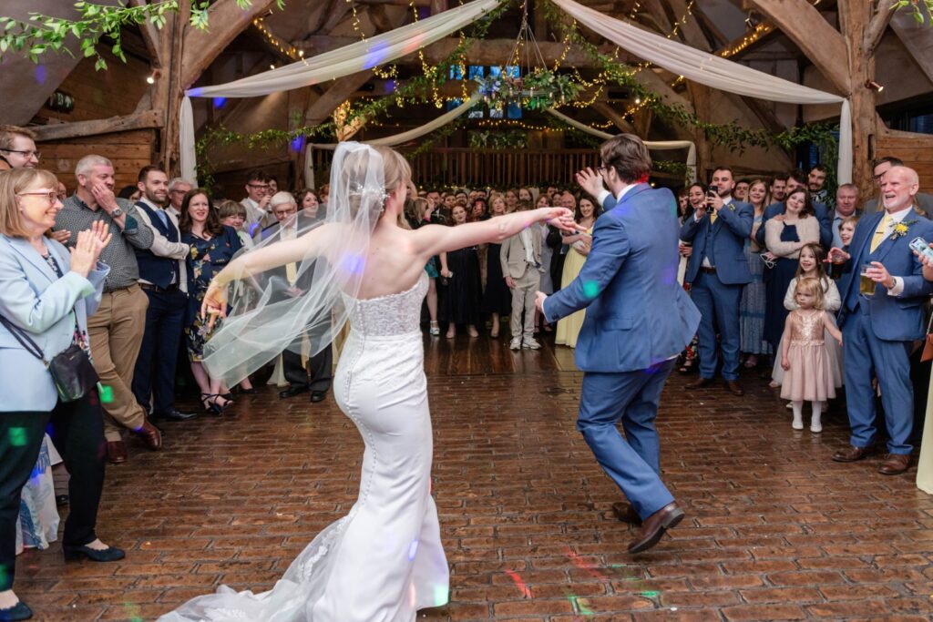 Bride and groom happily dancing with guests cheering around them at a rustic wedding venue.