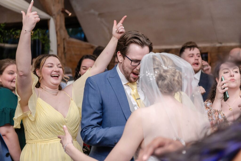 A group of guests, including a man in a blue suit and a woman in a yellow dress, are dancing and having a great time at a wedding reception.