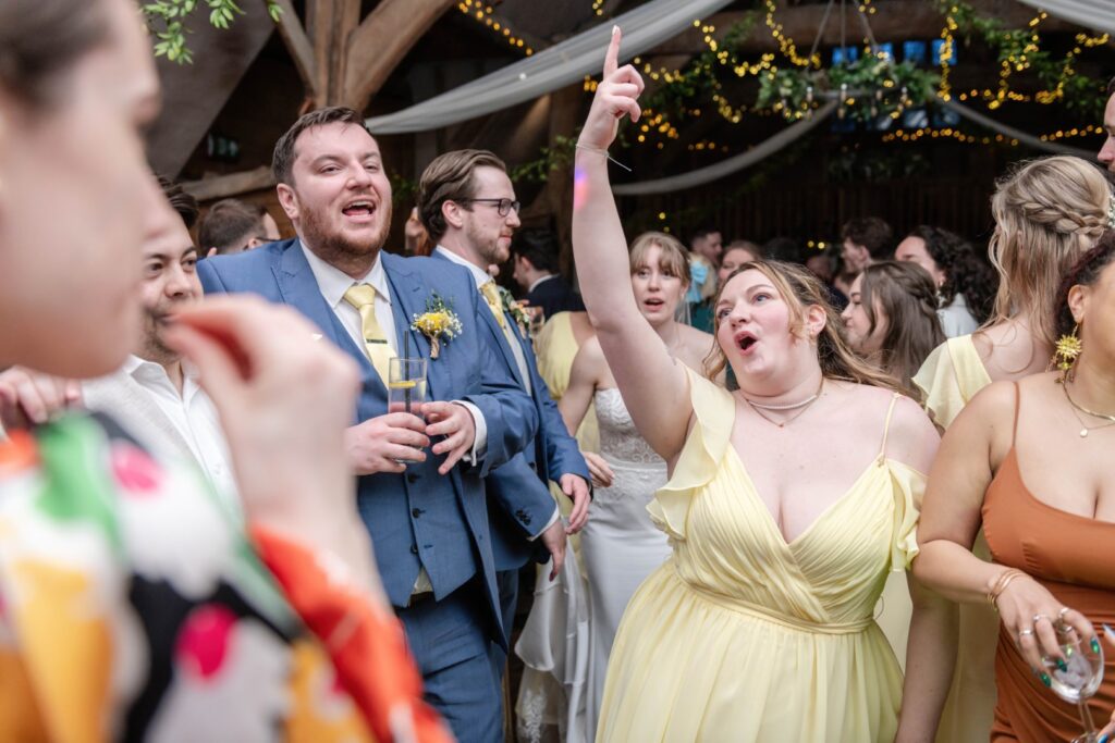 Guests at a wedding reception, including bridesmaids in yellow dresses, laughing and pointing upwards, with fairy lights in the background.