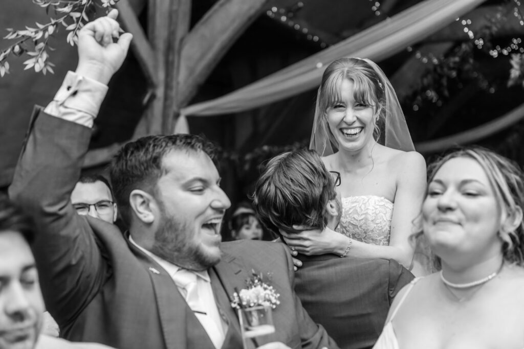 A black and white photograph capturing a moment of laughter and joy amongst a group of people at a wedding, with a bride smiling brightly in the center.