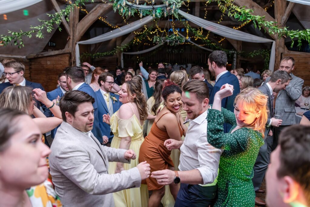 A group of elegantly dressed guests dancing and enjoying themselves at a festive wedding reception with string lights and greenery in the background.