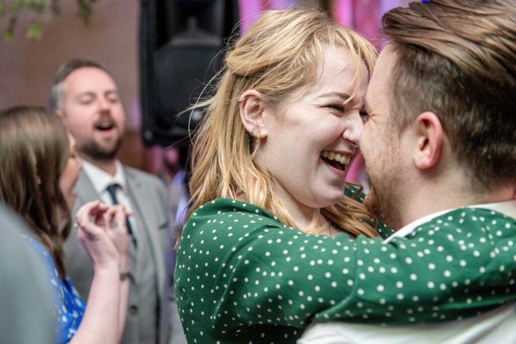 Two people sharing a heartfelt laugh while dancing closely at a social event, with others in the background.