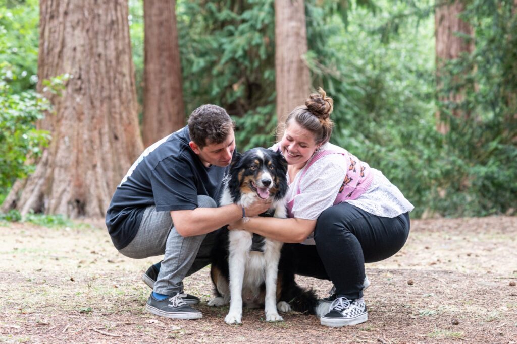 A couple smiling and hugging their black and white dog in a wooded park setting.