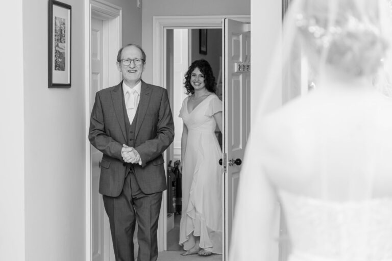 Black and white photo of a smiling man in a suit and a woman in a white dress standing in a doorway, with the focus on the bride's back in the foreground.