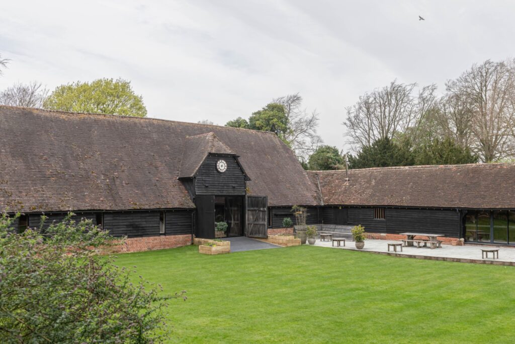 Lains Barn. A renovated black barn with clock, surrounded by a lush green lawn and trees under a cloudy sky.