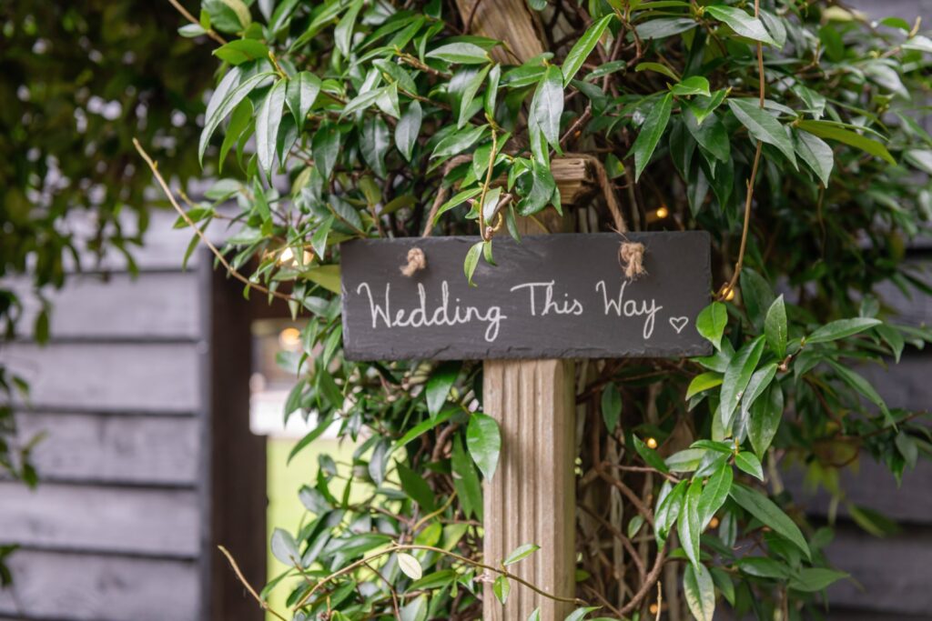 A blackboard sign with white lettering hanging on a post amidst green leaves that reads "Wedding This Way" with a heart symbol.