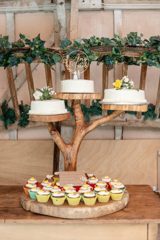 A rustic wedding cake stand with two cakes and an assortment of cupcakes on wooden slabs, garnished with green foliage and flowers, and a personalized cake topper with the names Amy & Jacob.