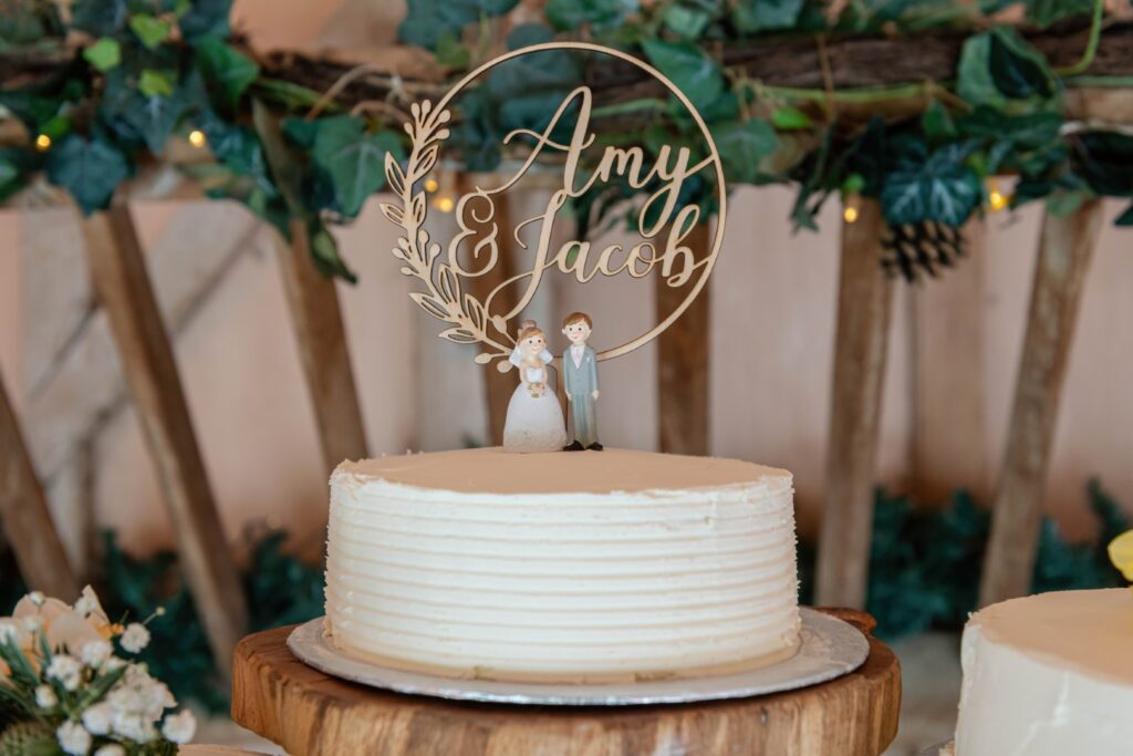 A simple white frosted wedding cake on a wooden stand with a personalized 'Amy & Jacob' cake topper, flanked by decorative greenery and fairy lights.