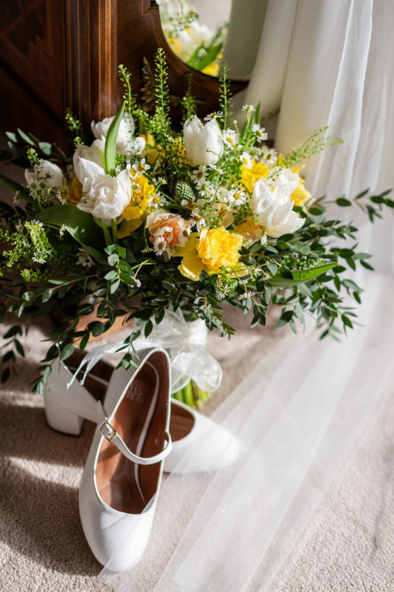 A pair of white bridal shoes placed beside a vibrant wedding bouquet with white and yellow flowers on a carpeted floor.