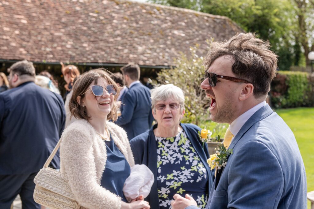 A woman and a man laugh heartily while conversing in a lively outdoor event with guests in the background.