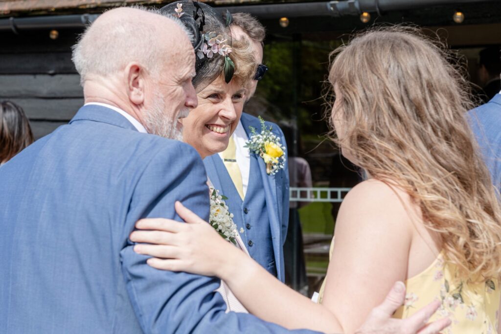 A close-up of three people at a wedding reception, with a man and a woman in the center smiling while embracing another woman, all dressed in formal wear with wedding flowers.