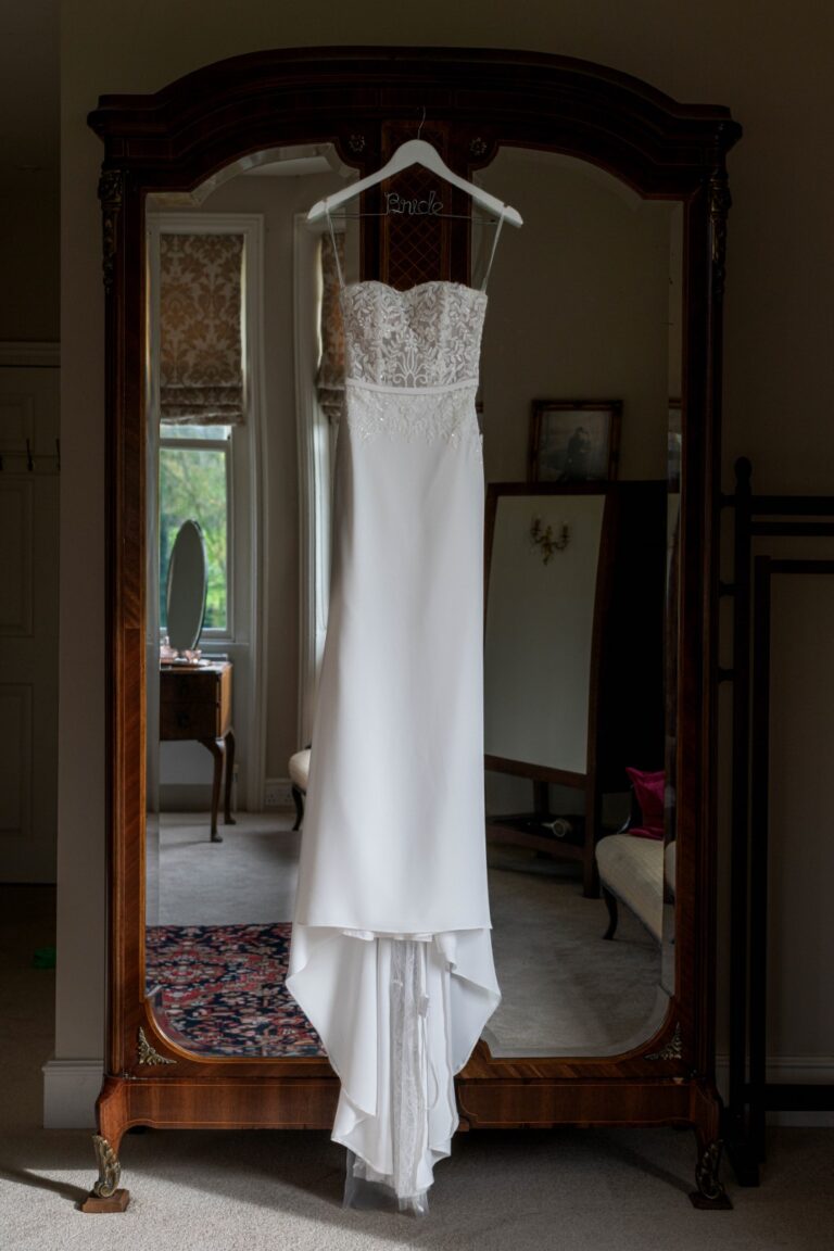 A white wedding dress hanging on a personalized hanger, reflected in an antique full-length mirror in a room with classic decor.