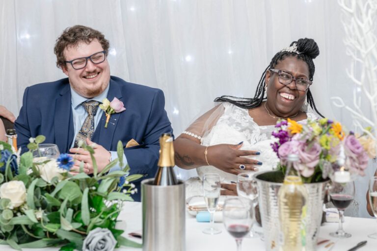 A laughing couple seated at a wedding reception table, surrounded by floral decorations, champagne, and glasses.