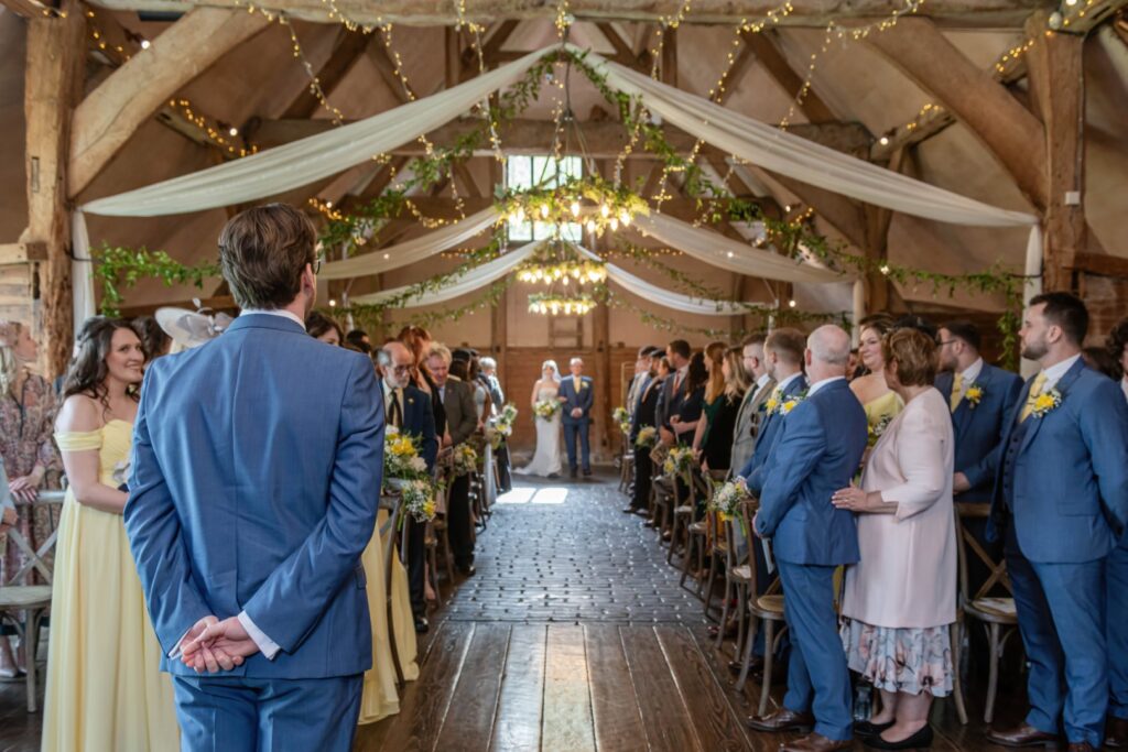 A wedding ceremony inside a rustic barn with wooden beams, guests lined up along the aisle, and a groom waiting at the altar.