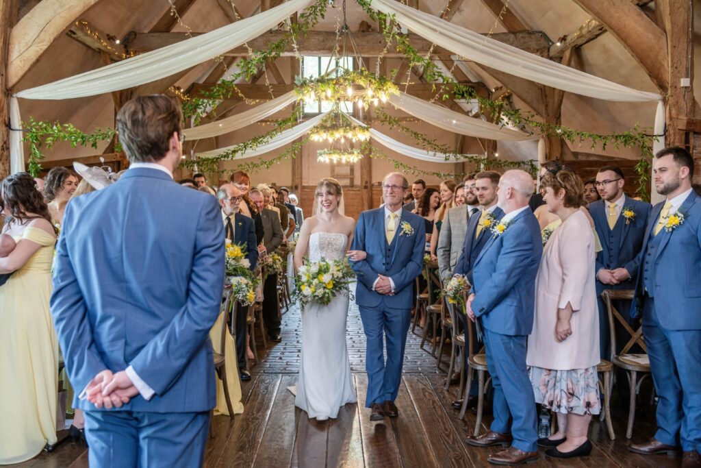 A bride walking down the aisle with her father, smiling at guests, in a warmly lit barn decorated with white drapes and greenery.