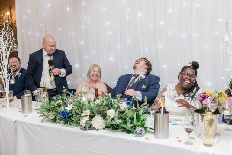 Guests at a wedding reception laughing joyously while a man speaks into a microphone, set against a backdrop of twinkling lights.