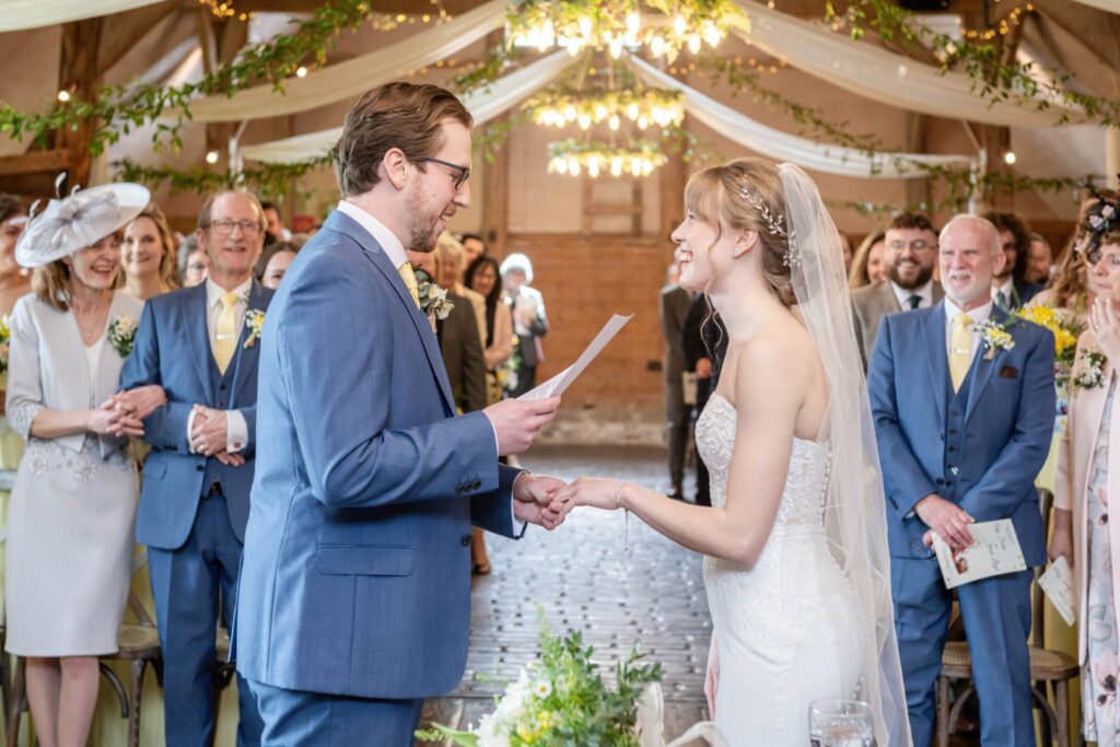 A bride and groom exchanging wedding vows in a beautifully decorated venue with smiling guests in the background.