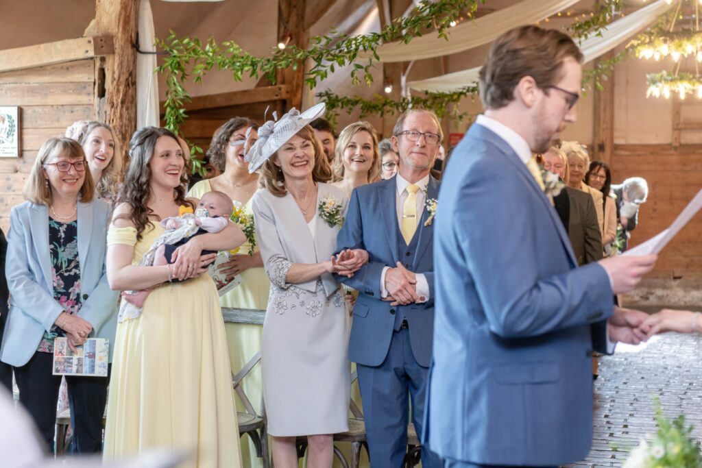 A group of elegantly dressed guests smiling and paying attention to a wedding speech, with a focus on a joyful couple probably the parents of the bride or groom.