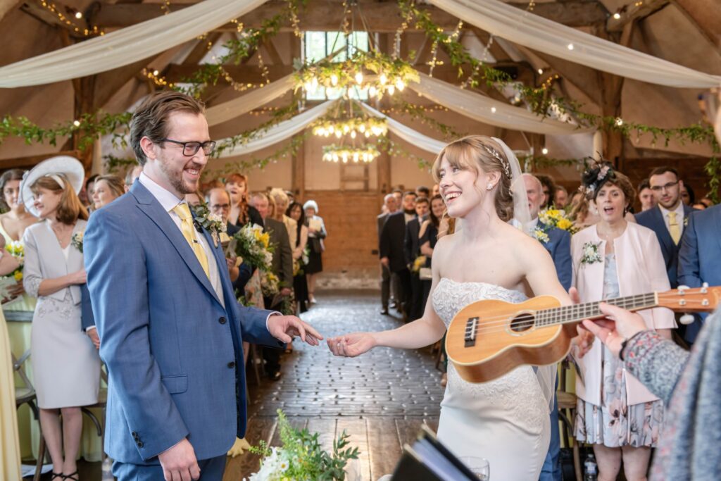 Bride and groom smiling at each other during a wedding ceremony in a barn decorated with fairy lights and drapery, with the bride holding a ukulele and guests looking on.