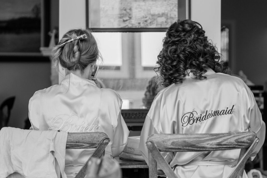 Two women in satin robes with "Bride" and "Bridesmaid" written on the back, sitting and facing away from the camera in a black and white photo.