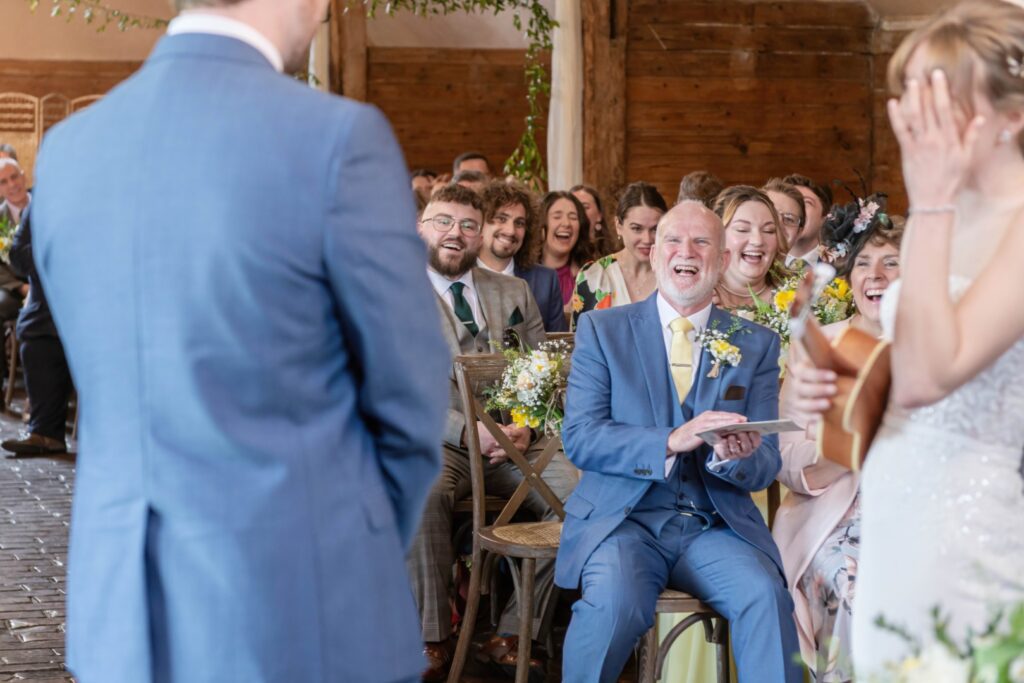 A group of wedding guests laughing joyously while seated during a ceremony, with focus on an older man in a suit clapping and laughing.