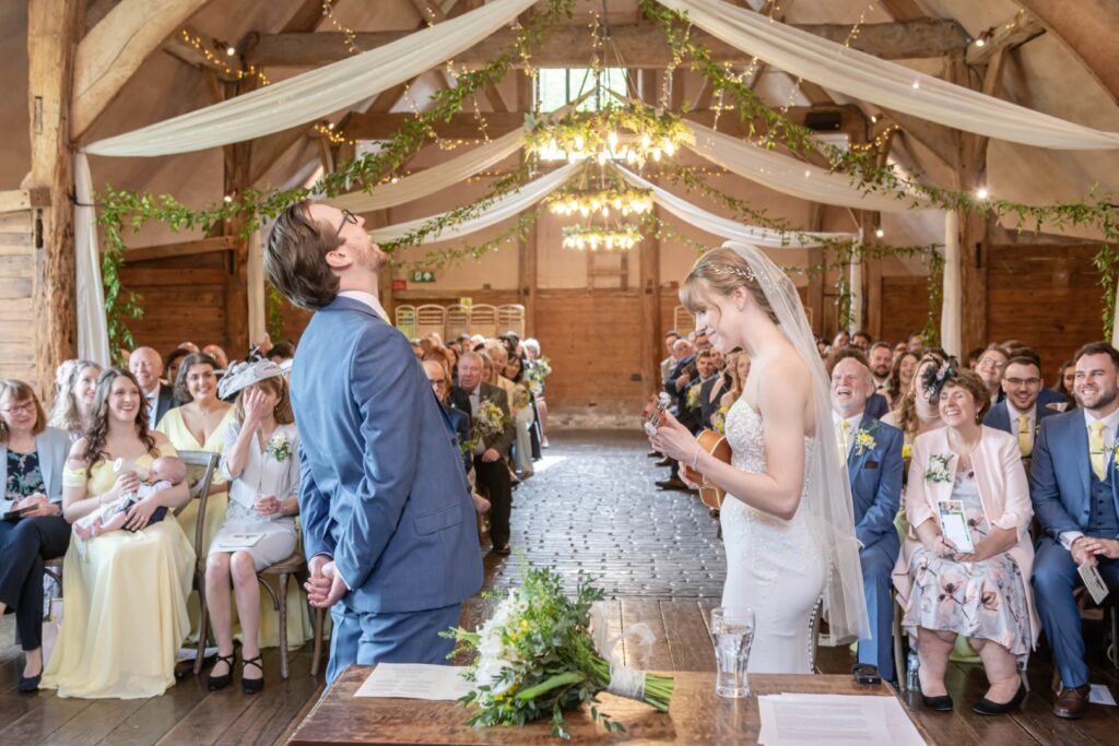 A bride playing a ukulele and laughing with the groom who is looking up and laughing as well, surrounded by guests in a rustic wedding venue with exposed wooden beams and string lights.