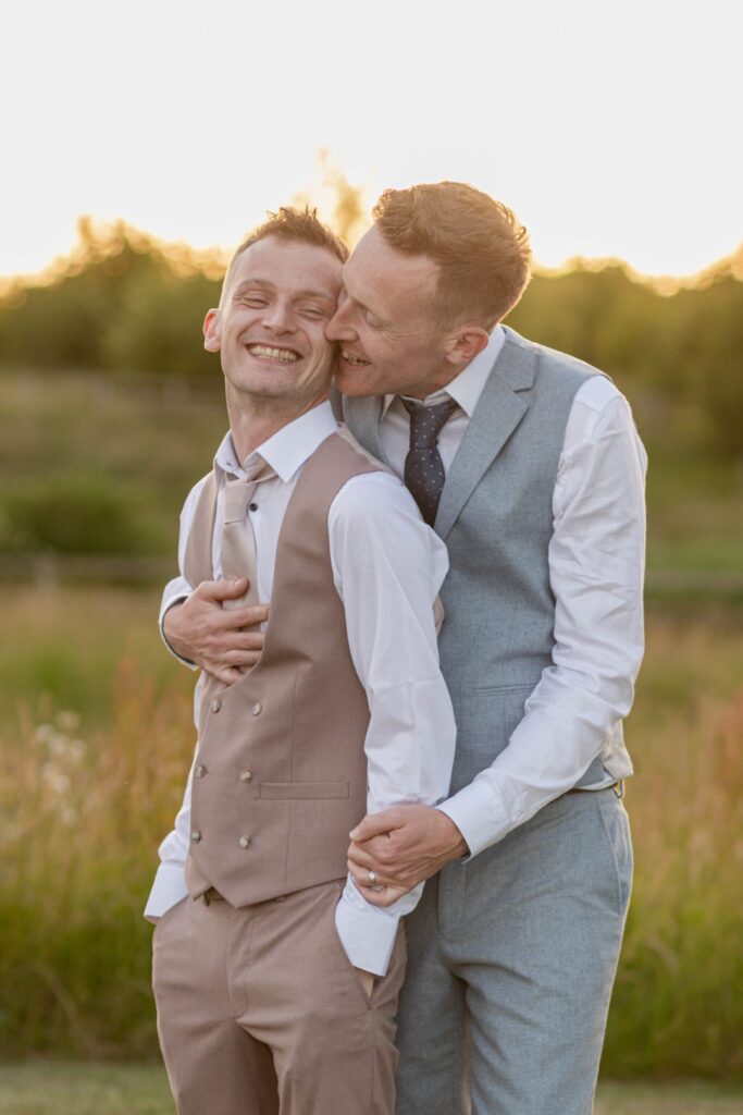 Two men hugging and smiling in a field during sunset, both dressed in smart vests and trousers.