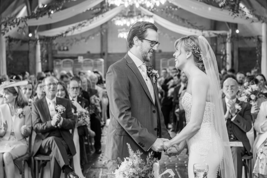 A black and white photo of a bride and groom holding hands and exchanging vows during a wedding ceremony with guests looking on.