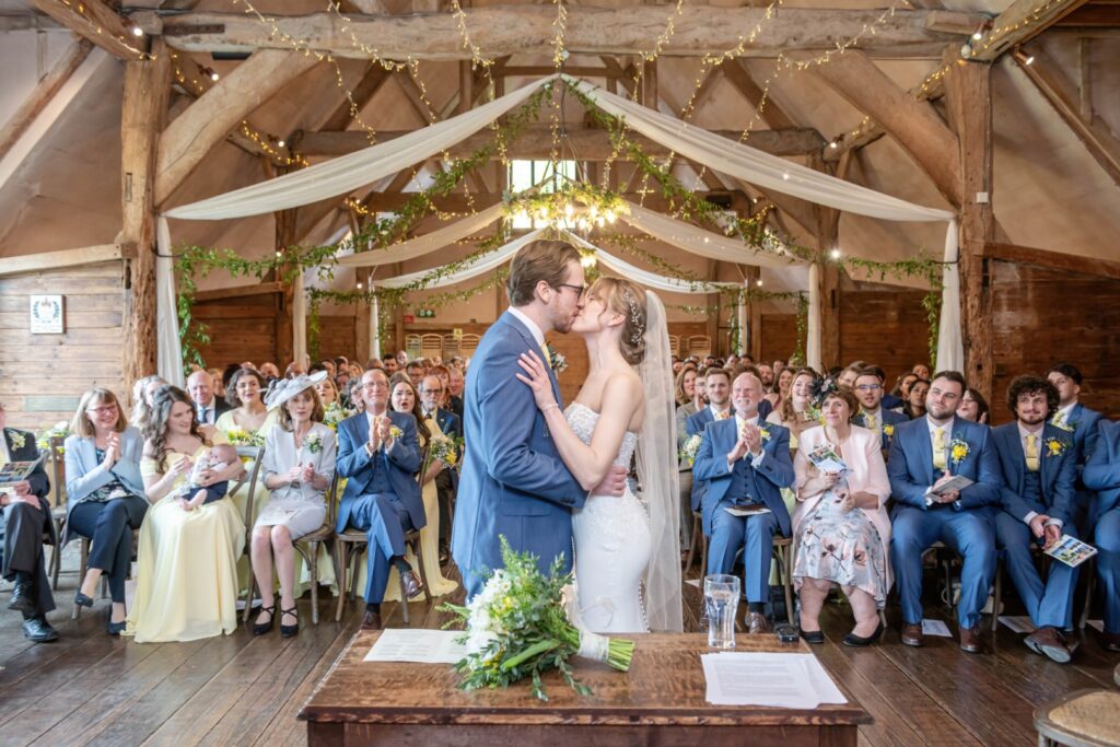 A bride and groom kissing in front of seated guests at a rustic barn wedding venue, with wooden beams and string lights overhead.