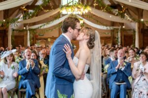 Bride and groom kissing at the altar in a rustic wedding hall with guests applauding in the background.