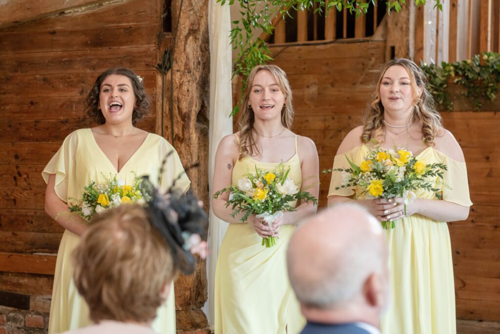 Three bridesmaids in yellow dresses holding flower bouquets smiling during a wedding ceremony inside a rustic venue.