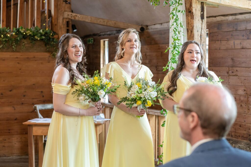 Three bridesmaids wearing yellow dresses holding bouquets in a rustic wedding setting, smiling and standing in a row with wooden interior in the background.