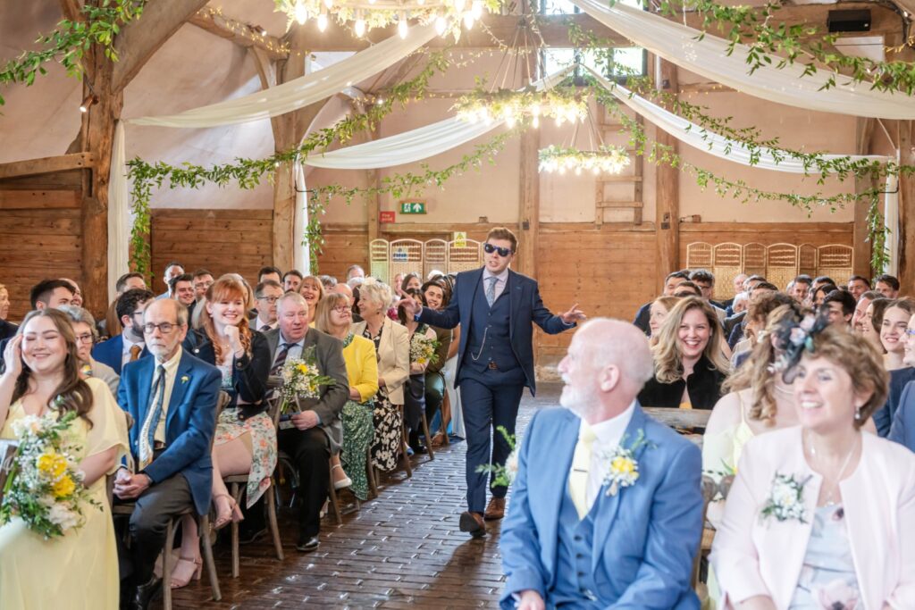 A groom in sunglasses walks down the aisle with a cheerful expression as wedding guests look on, seated inside a rustic venue decorated with white drapes and greenery.