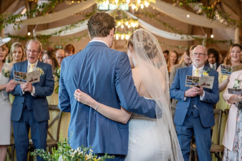 A bride and groom share an embrace during their wedding ceremony, with guests looking on in the background.