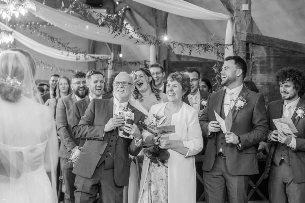 Black and white photo of guests laughing and smiling joyfully at a wedding ceremony with a bride in the foreground facing away.